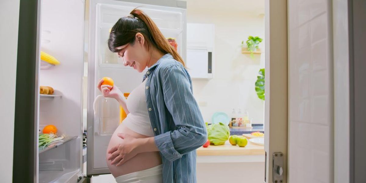 Várandós nő a nyitott hűtőszekrény előtt, egy narancsot a kezében tartva áll