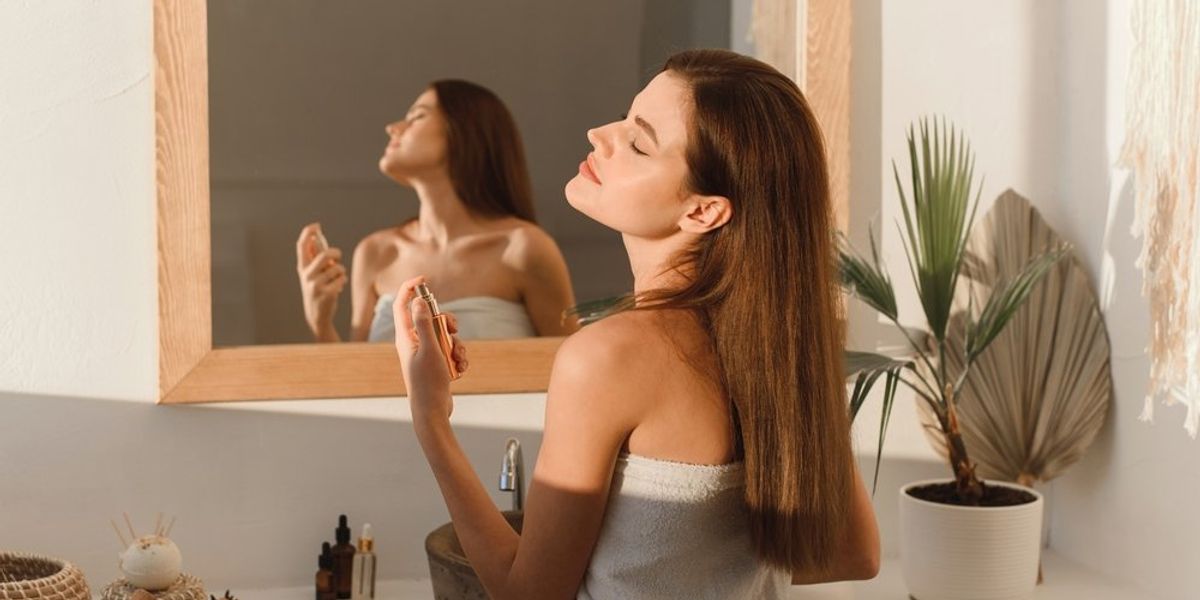 Fiatal, barna hajú nő parfümmel fújja be magát tükör előtt ülve