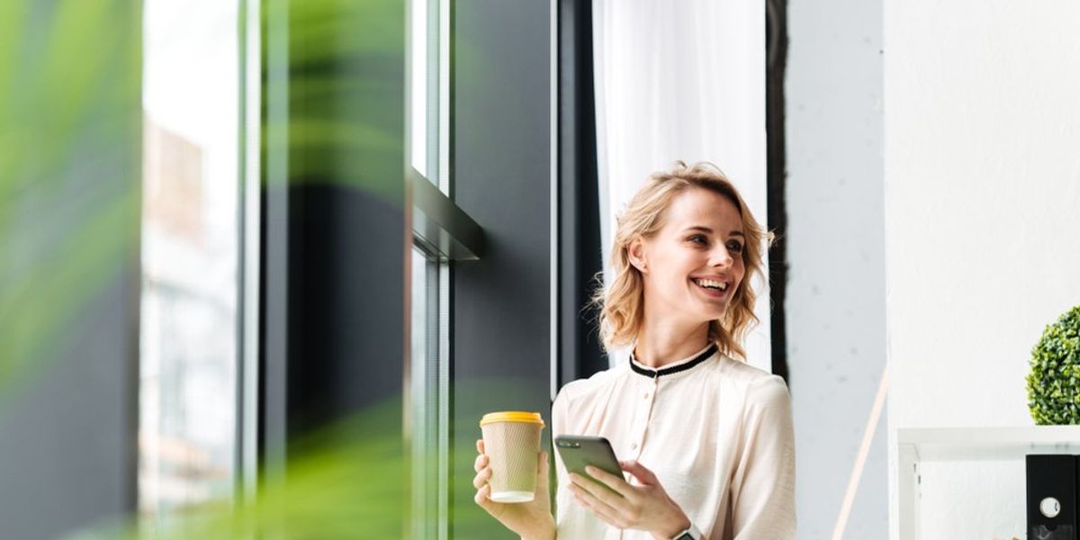 egy nő áll az ablaknál az irodában egy kávéval a kezében