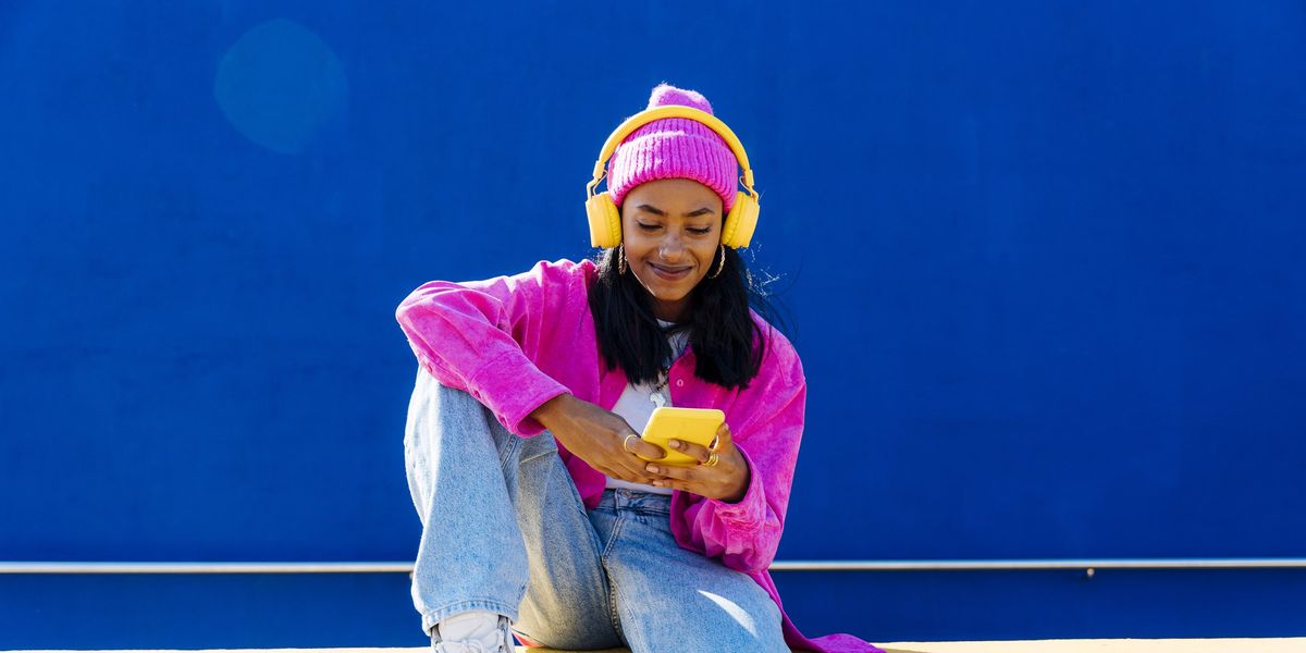 mosolyogva zenét hallgat egy lány sárga fejhallhatón keresztül, egy sárga falon ülve