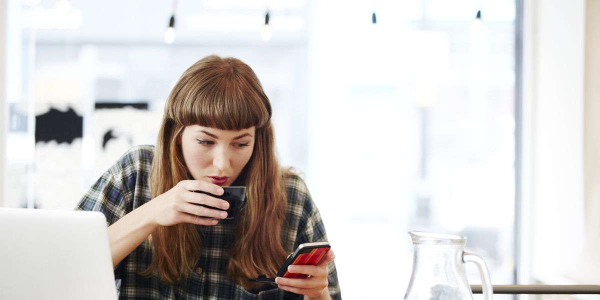 egy nő kávét iszik a laptopja előtt, miközben a telefonját nézi