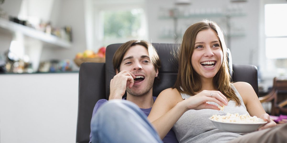 egy pár nevetve popcornt eszik és filmet néz egy fotelban ülve