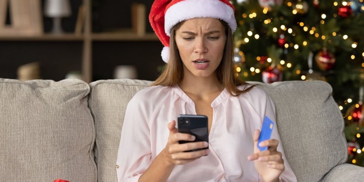 egy mikulás sapkás nő idegesen nézi a telefonját a bankkártyájával a kezében, mögötte karácsonyfa