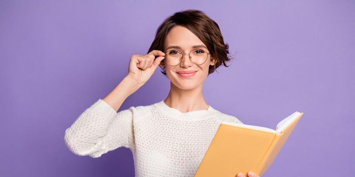 Nő fehér pulcsiban, könyvvel a kezében, szemüvegben, lila háttér előtt