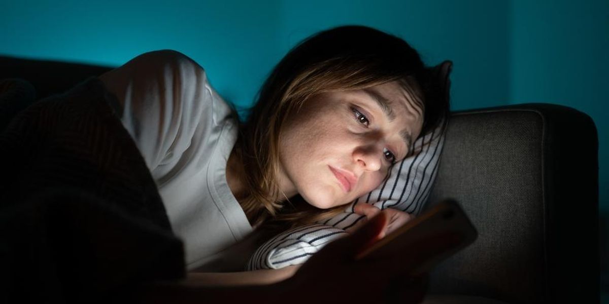 Szomorú nő fekszik az ágyban, és a mobilján nézegeti a volt barátját a közösségi médiában