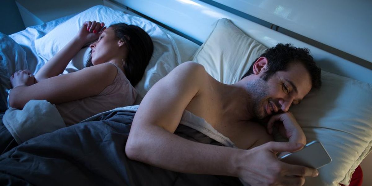 Nő és férfi egymásnak háttal fekszik az ágyban, a férfi telefonozik