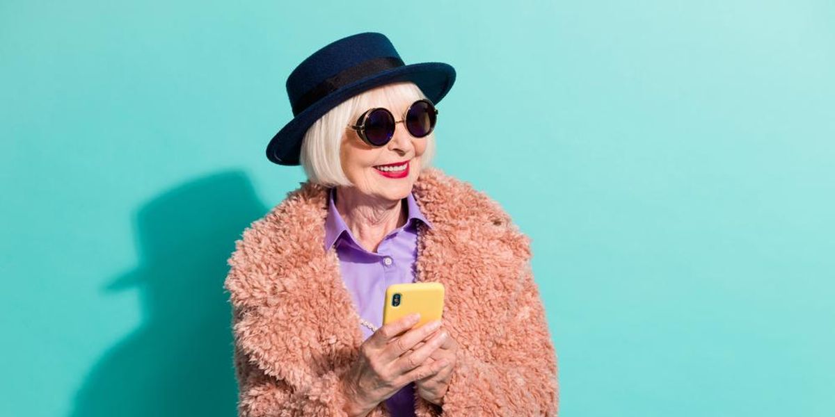 Ősz hajú nő kalapban, napszemüvegben és rózsaszín kabátban telefonozik