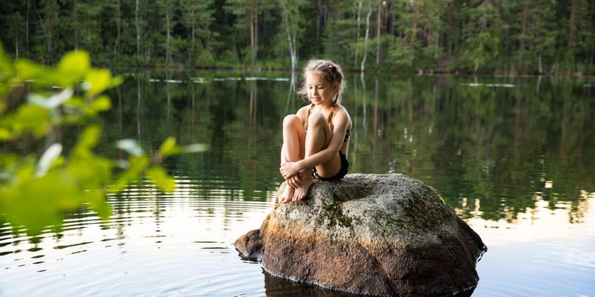 kislány ül egy nagy kövön a víz közepén, kacsa úszik mellette