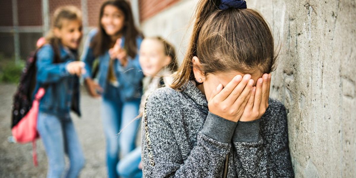 egy kislány a tenyerébe temeti az arcát, mögötte gúnyolódik rajta a többi gyerek