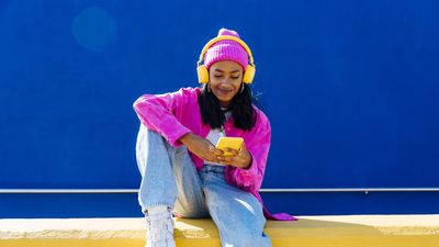 mosolyogva zenét hallgat egy lány sárga fejhallhatón keresztül, egy sárga falon ülve