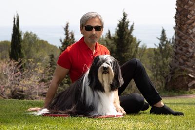Náray Tamás piros pólóban és napszemüvegben ül a kutyájával a fűben