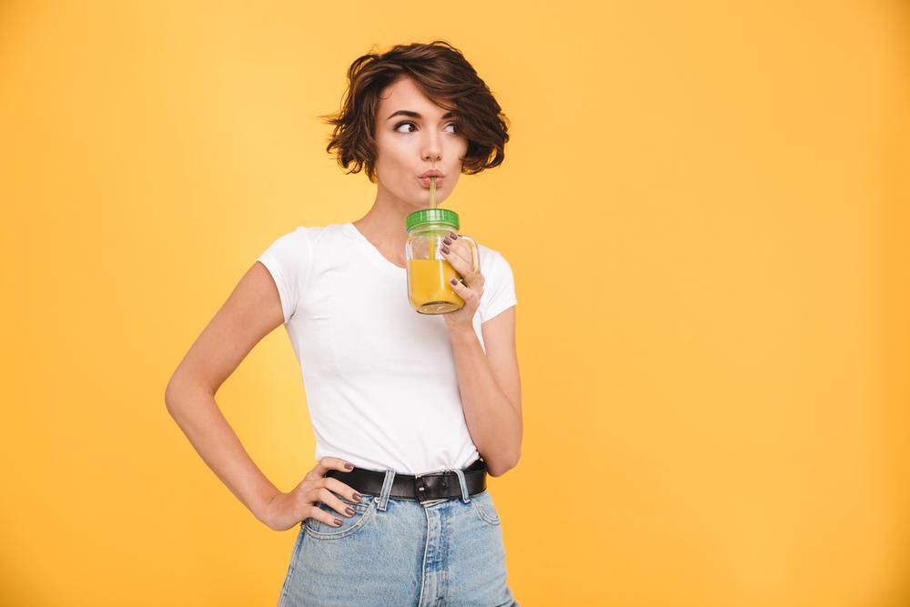 Fiatal nő narancslevet iszik egy pohárból, narancssárga háttér előtt