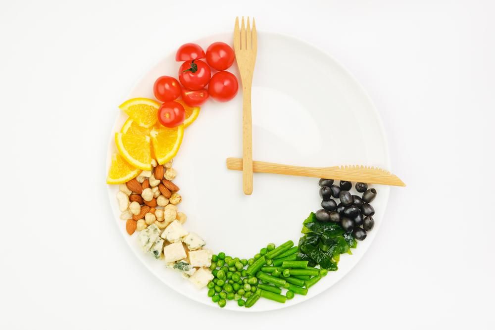 Színes ételek és evőeszközök óra alakban elrendezve egy tányéron