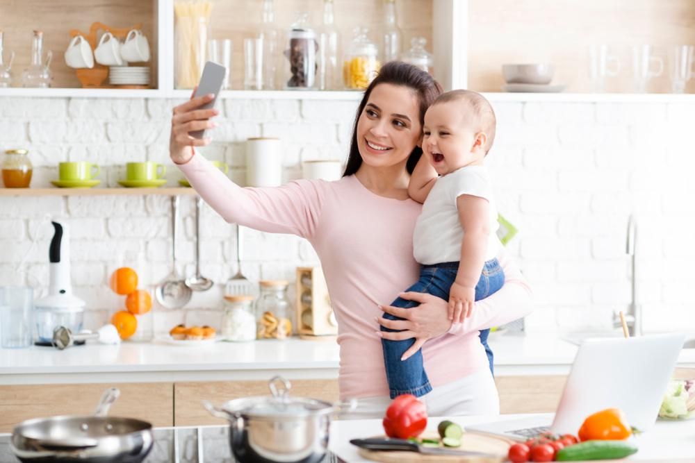 Nő a gyerekét fogja a konyhában és közben szelfizik