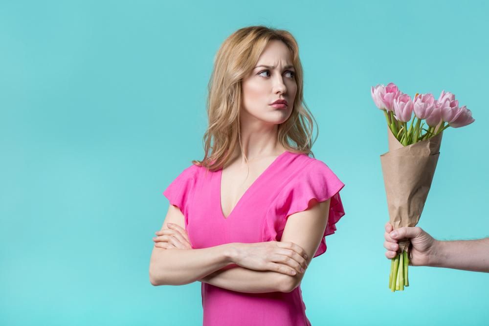 Szőke nő mérgesen néz, rózsaszín pólóban,  egy kéz tulipán csokrot nyújt neki
