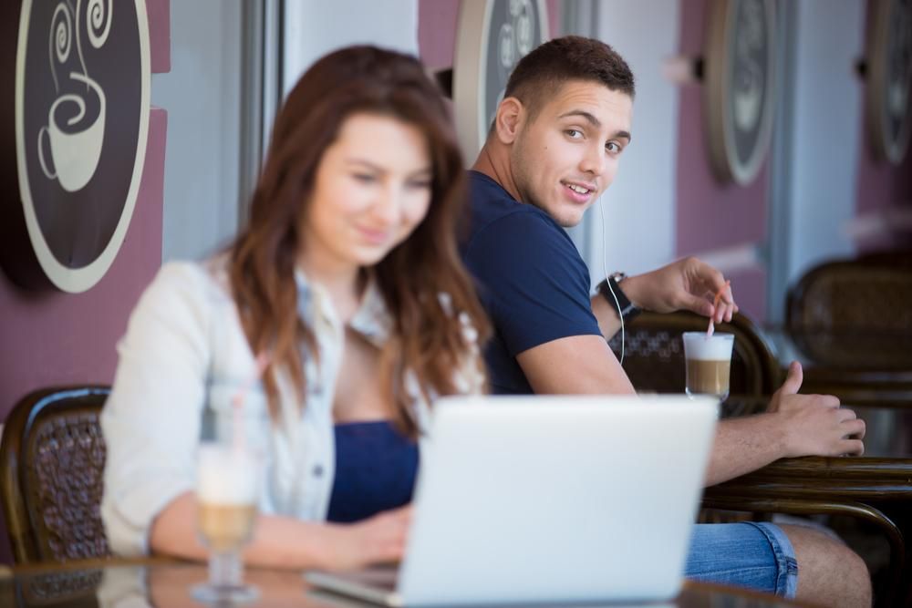 Barna hajú nő laptop előtt ül egy kávézóban, egy férfi nézi