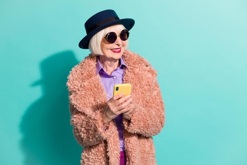 Ősz hajú nő kalapban, napszemüvegben és rózsaszín kabátban telefonozik