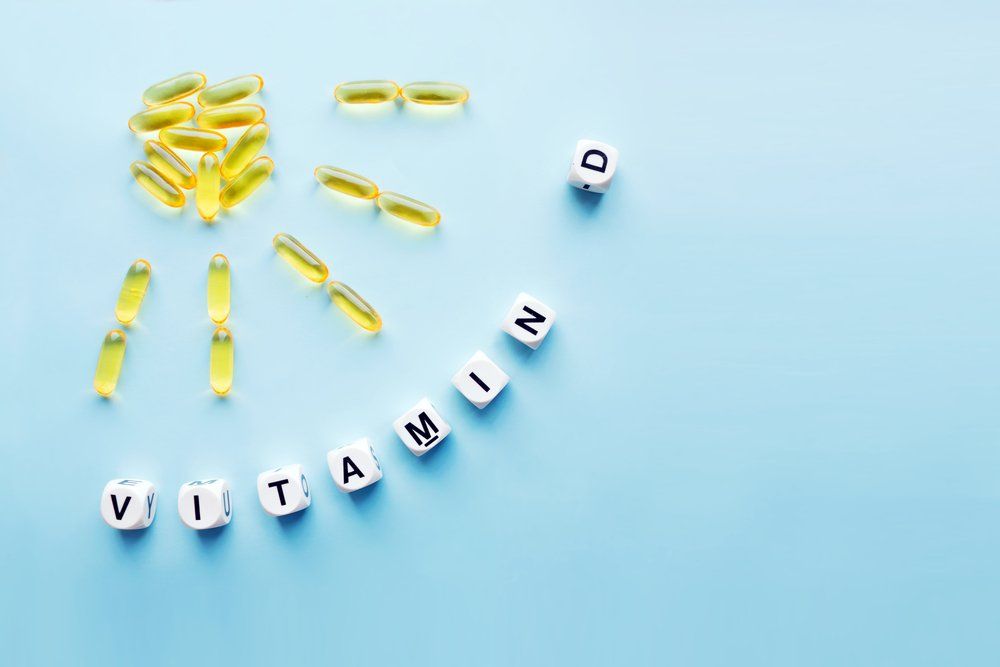 Sárga kapszulákból kirakott Nap forma, alatt D-vitamin szó fehér kockákon lévő betűkkel kirakva, kék háttéren